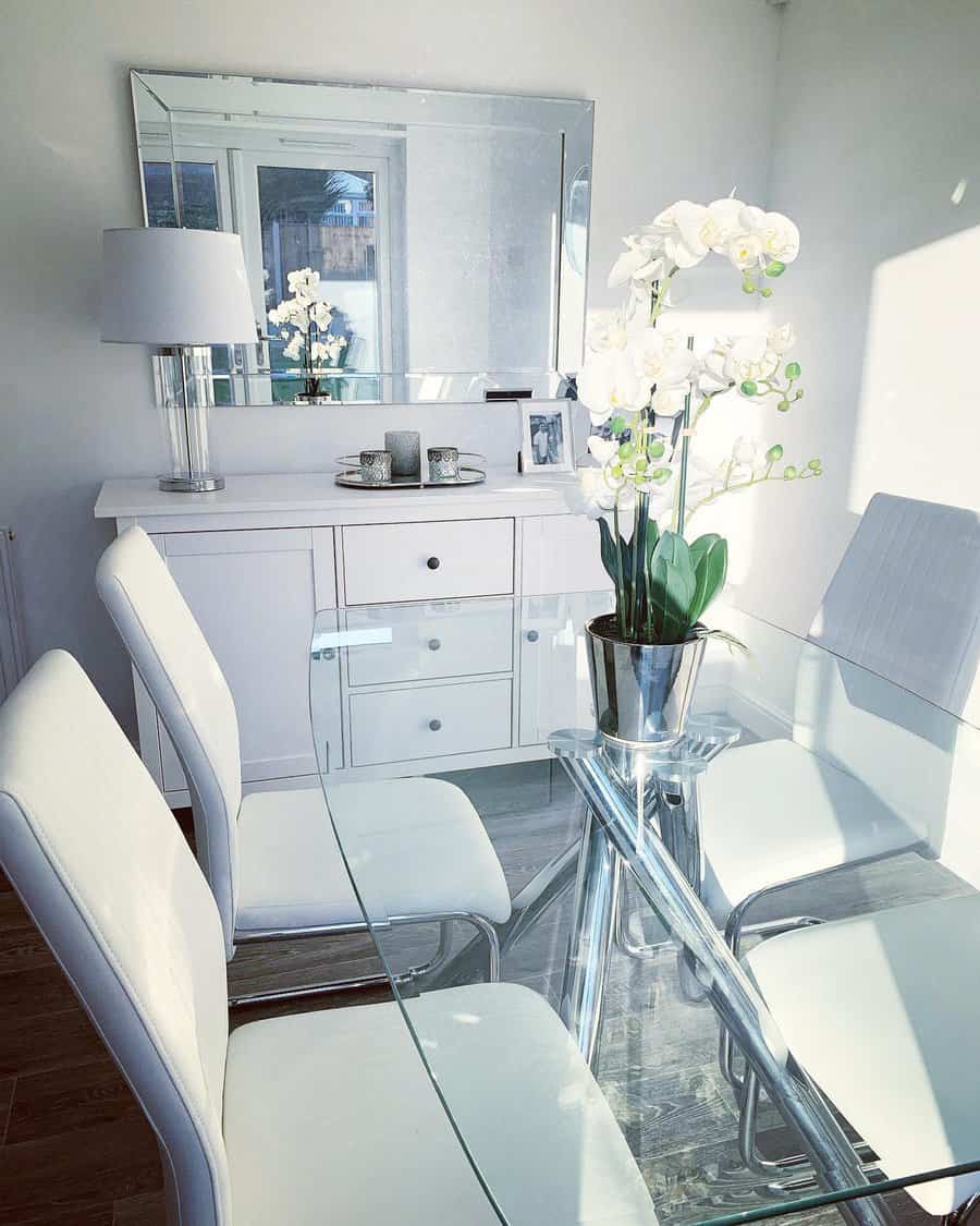 Modern dining room with a glass table, white chairs, and a sleek orchid centerpiece in a silver pot, reflecting natural light