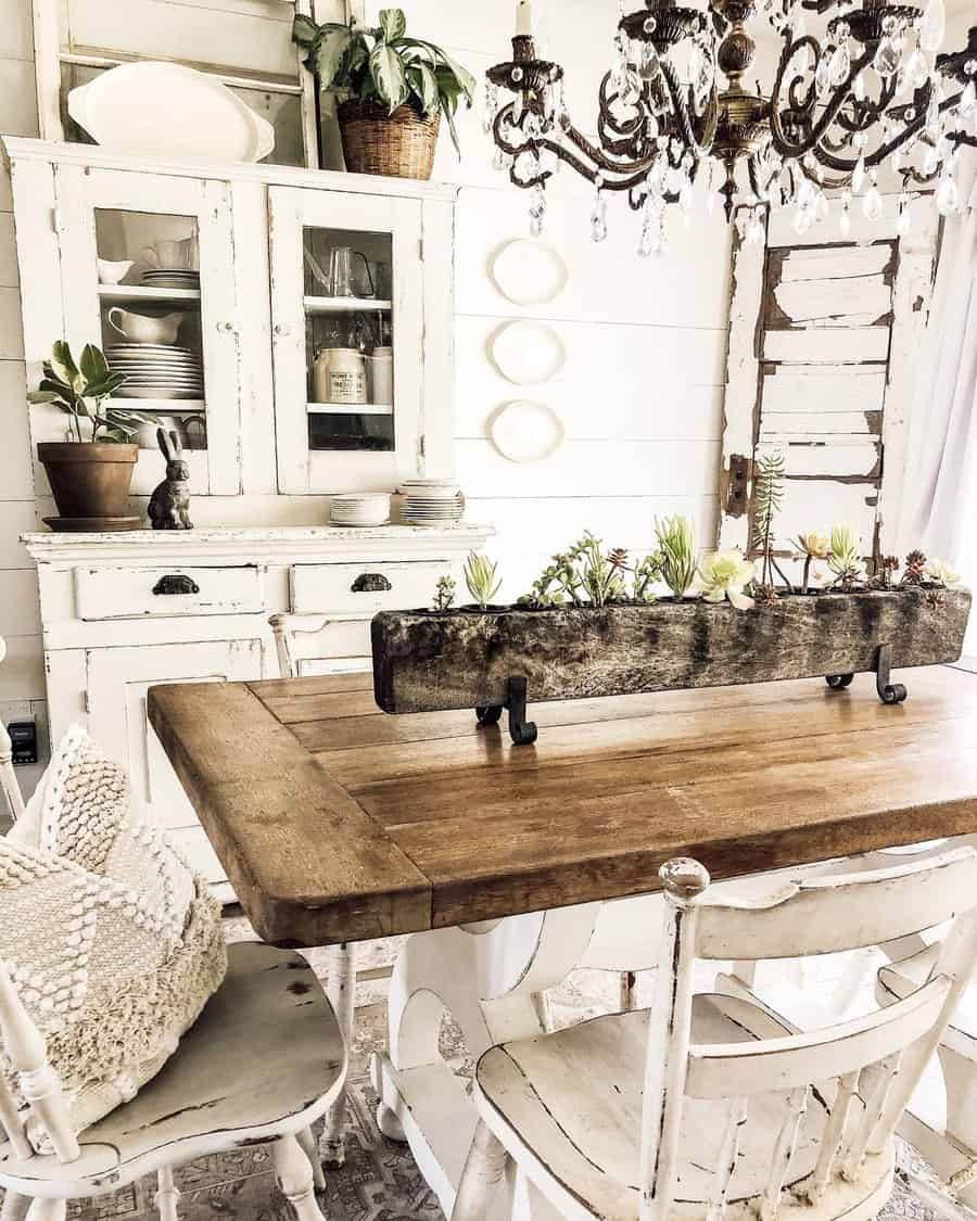 Rustic kitchen with a wooden table, white chairs, a white cabinet, and a chandelier featuring a table centerpiece with succulents