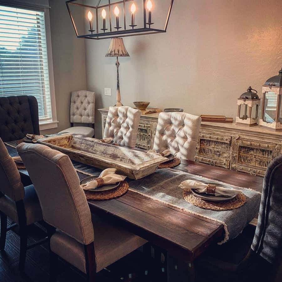 Rustic dining room with a dark wood table, tufted chairs, a distressed wooden tray centerpiece, woven placemats, and warm lighting