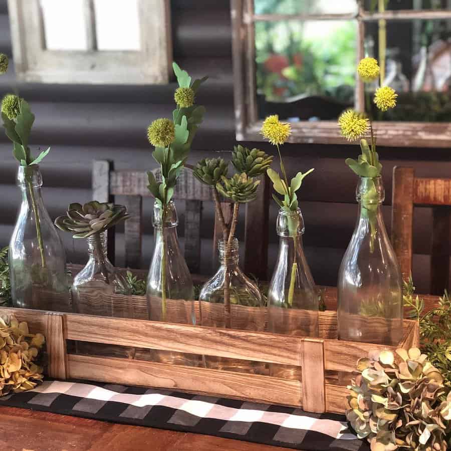 Glass bottles with green flowers and succulents arranged in a wooden crate on a table with a checkered cloth