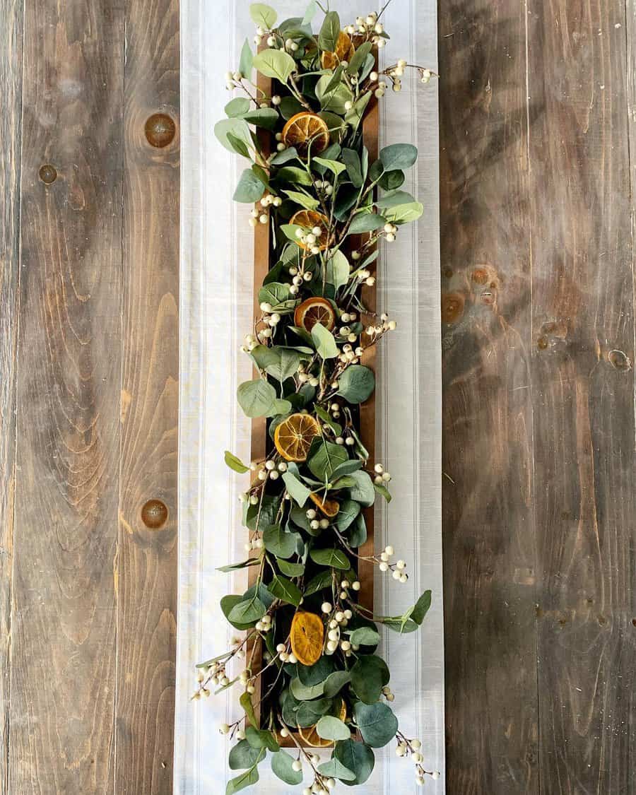 A wooden table centerpiece with eucalyptus leaves, dried orange slices, and white berries on a white runner