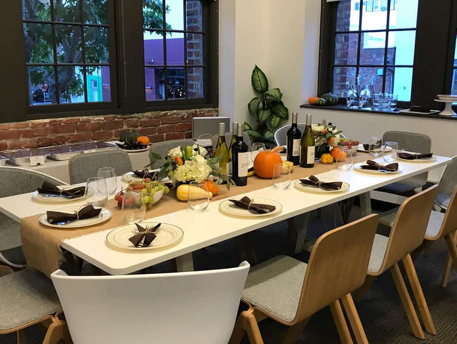 A festive dining table set with plates, wine, pumpkins, and autumn decorations, surrounded by modern chairs in a room with large windows