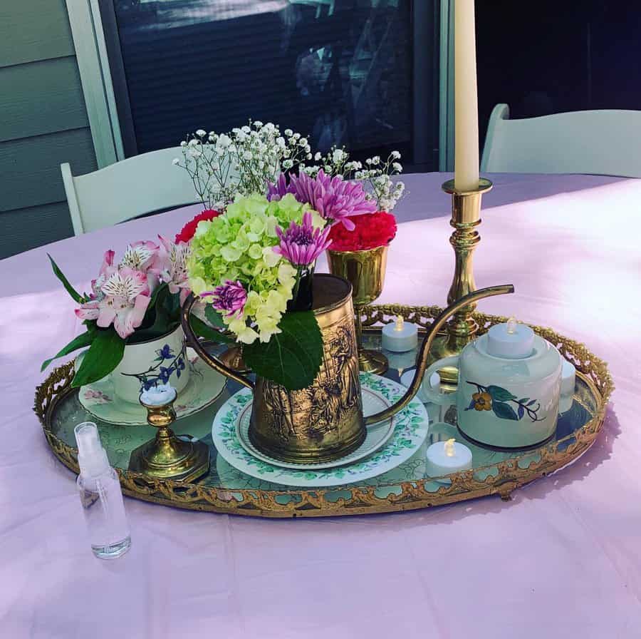 A decorative table centerpiece with flowers in a brass teapot, candles, china, and a tall candlestick on an ornate tray