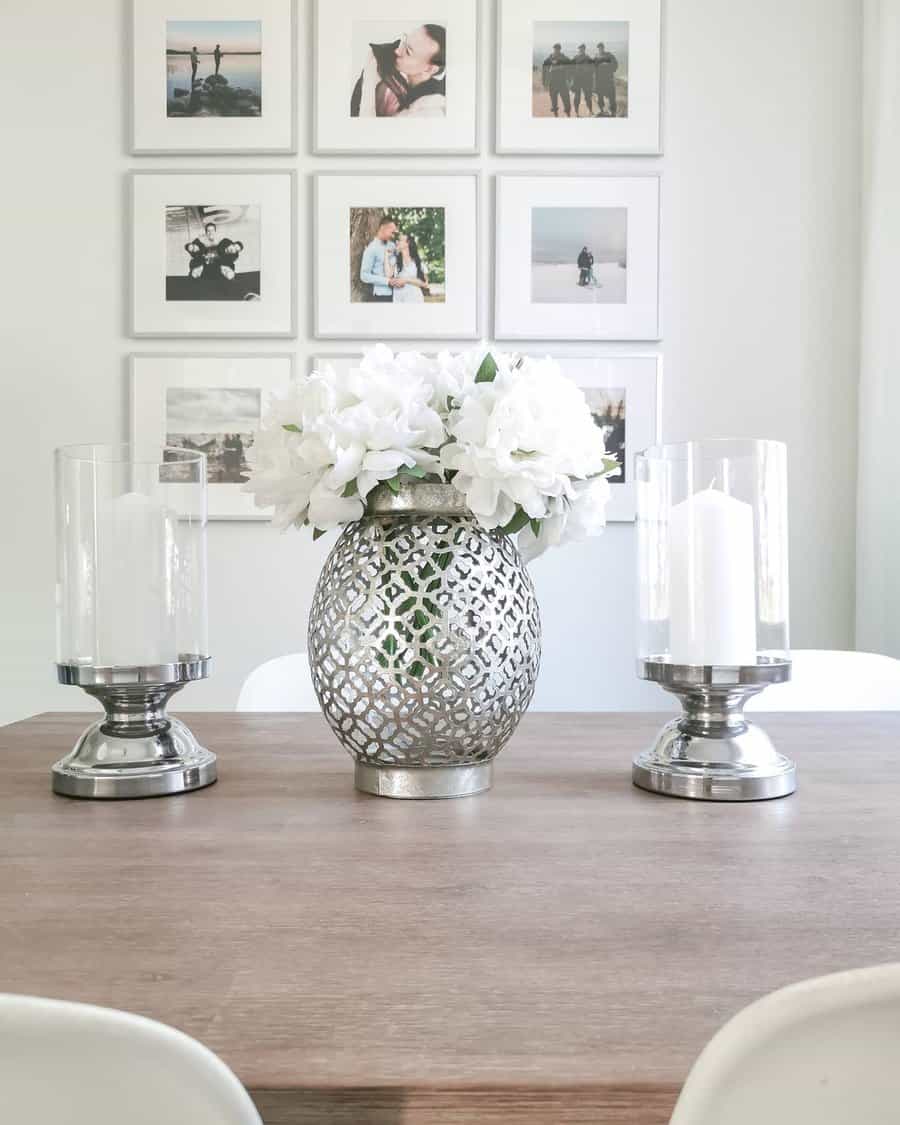 Decorative vase with white flowers on a wooden table, flanked by candle holders, with a gallery wall featuring photos in the background