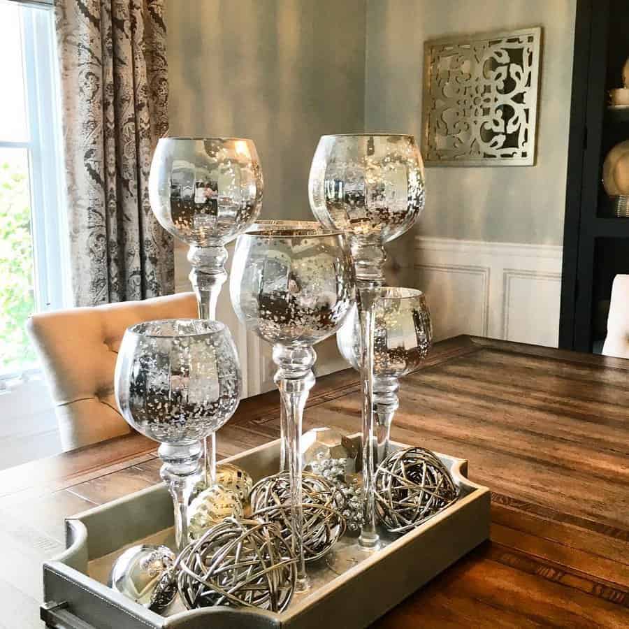 Decorative silver glass candle holders and metallic spheres on a wooden tray atop a dining table with ornate curtain backdrop