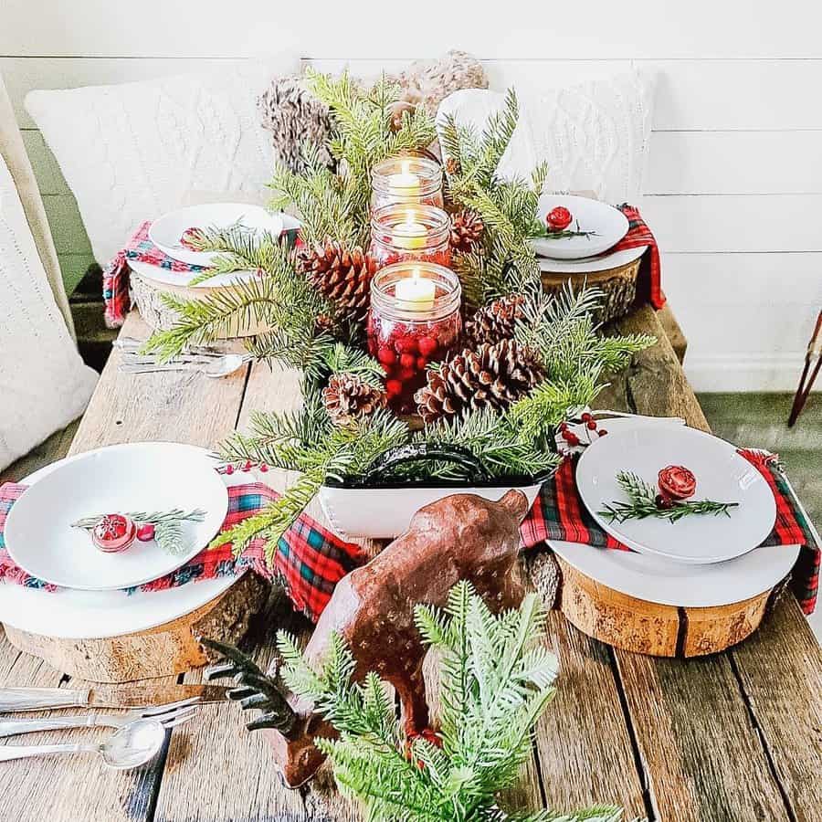 Rustic holiday table setting with pinecones, lit candles, plaid accents, wooden chargers, and evergreen branches on a wooden table