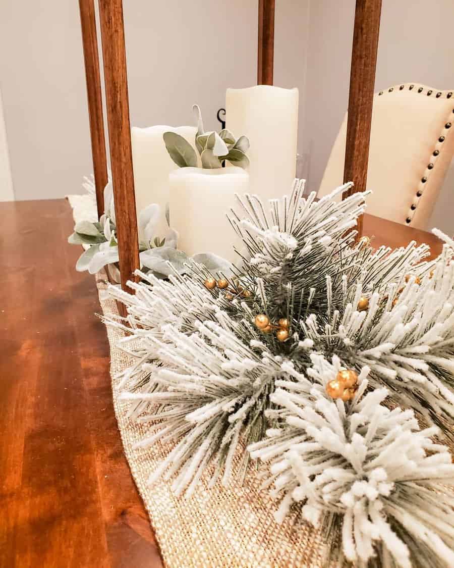 A table centerpiece featuring white candles, green pine branches with frosted tips, and a rustic wooden lantern on a fabric runner