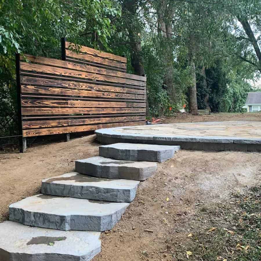 Backyard with a rustic wooden privacy screen, stone slab steps, and a circular stone patio, creating a secluded and natural outdoor retreat