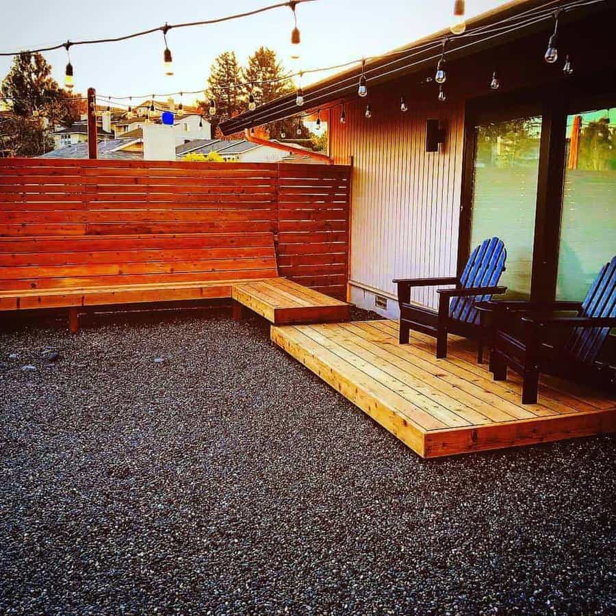 Modern backyard patio with a wooden slat privacy screen, built-in bench seating, string lights, and a raised wooden deck with Adirondack chairs for relaxation