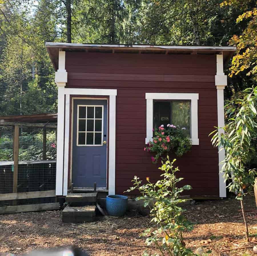 Small, red wooden shed with a blue door and window box with flowers, surrounded by trees and garden plants