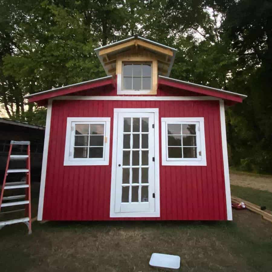 Small red shed with white trim and French doors, featuring a small loft window at the top, ladder and trees visible around