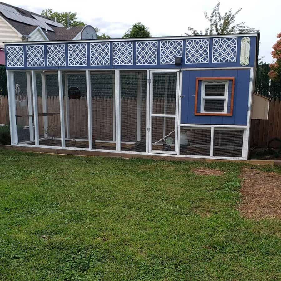 A blue chicken coop with a run, featuring lattice panels and a mesh enclosure, stands on a grassy yard