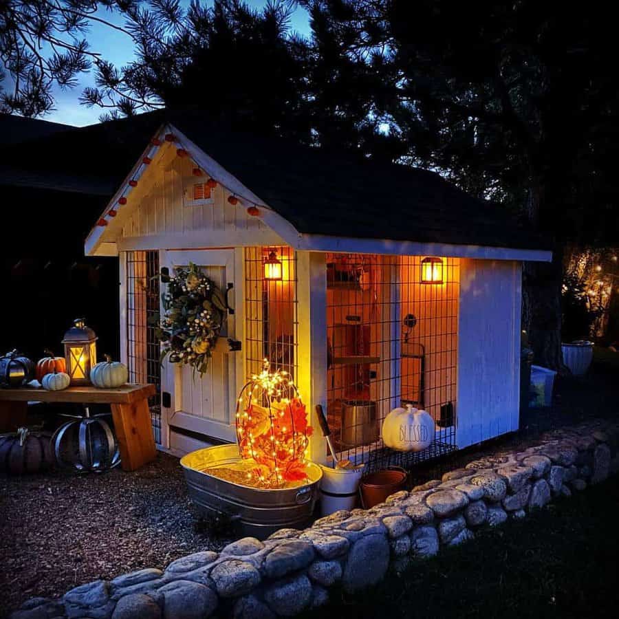 A cozy shed decorated with pumpkins and a lit wreath, surrounded by a stone border, warmly illuminated at dusk