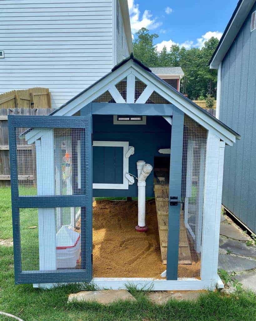 A chicken coop with a slanted roof, wire mesh doors, a sand-covered floor, and a nesting box, situated in a backyard