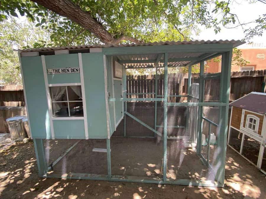 A blue chicken coop labeled "The Hen Den" under a tree with a fenced enclosure, beside a small wooden house, in a sunlit yard