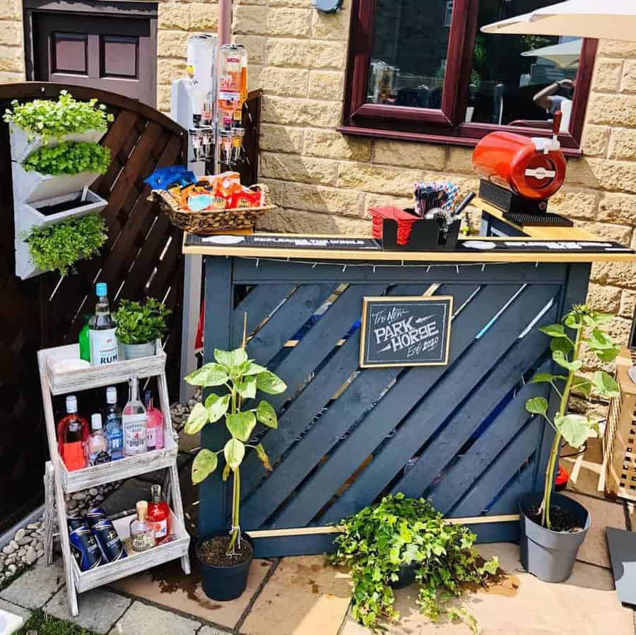 Outdoor pallet bar with a rustic design, featuring a stocked liquor shelf, mounted drink dispensers, and potted plants for decor