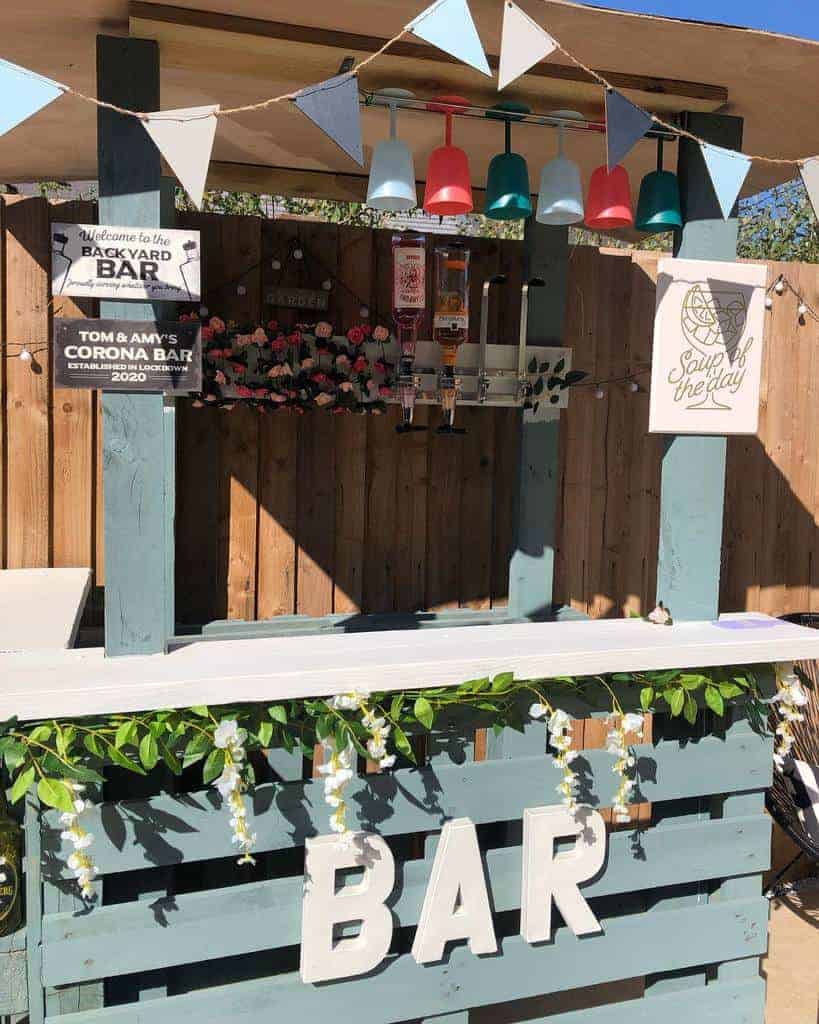 Rustic backyard pallet bar with bunting, hanging glasses, liquor dispensers, floral garland, and personalized signage for a cozy setup