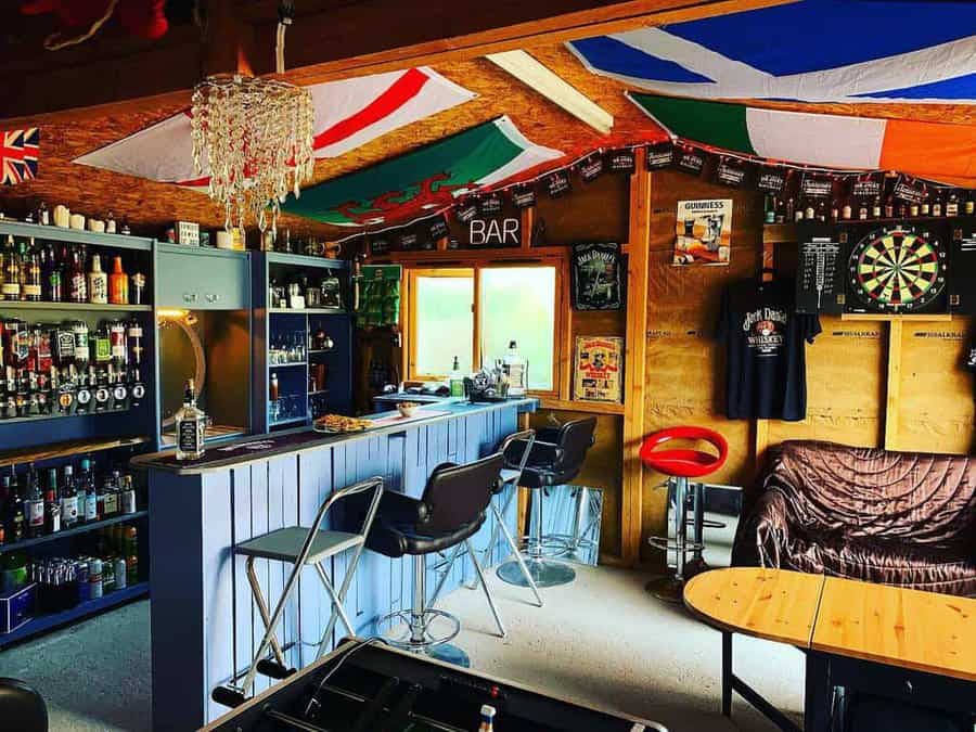 Cozy home bar with a rustic pallet counter, stocked liquor shelves, dartboard, flags, and pub-style seating for an inviting atmosphere