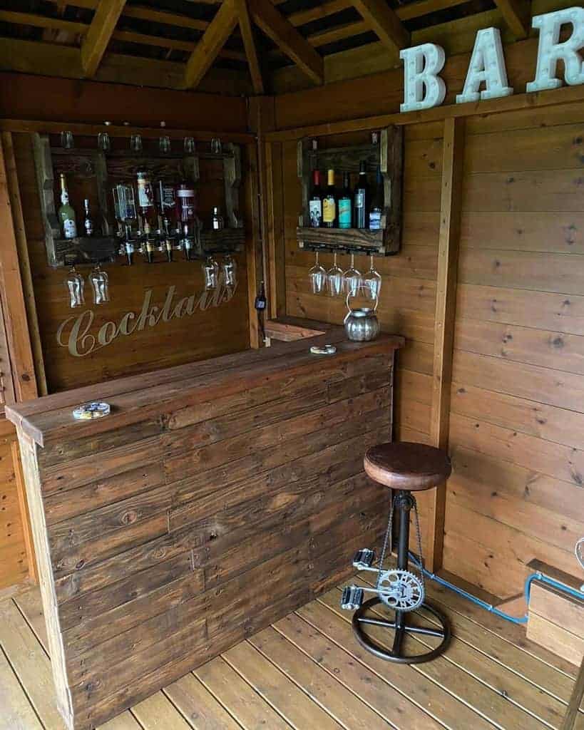 Rustic wooden pallet bar with wall-mounted liquor shelves, hanging glass storage, and a unique bicycle pedal barstool for a vintage touch