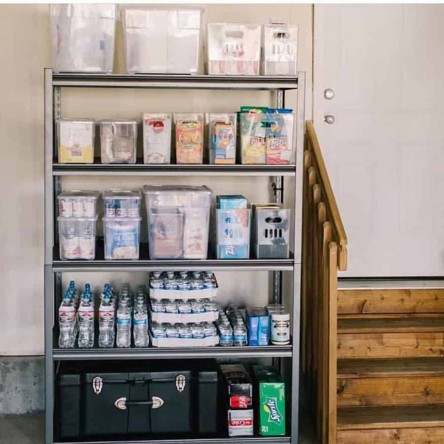 A metal shelving unit with organized supplies, including water bottles, canned goods, and plastic containers, next to wooden stairs