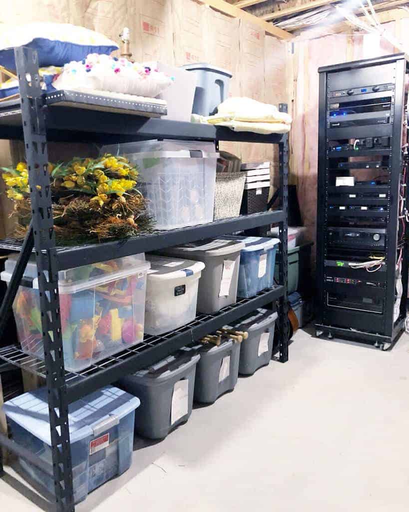 Shelving with storage bins and decor items next to a tall server rack in a basement room