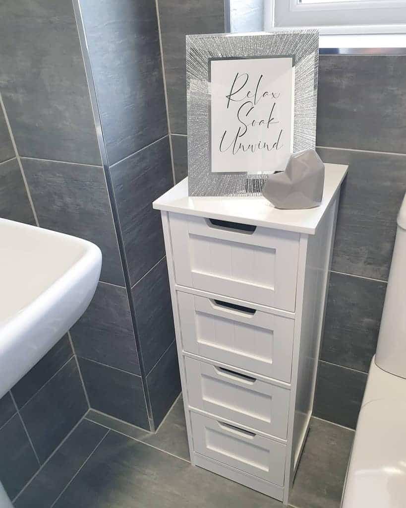 A white four-drawer cabinet in a bathroom corner with a framed sign reading "Relax Soak Unwind" and a decorative rock on top