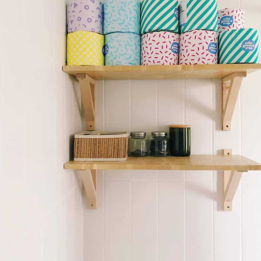 Wooden shelves with colorful toilet paper rolls on the top shelf and jars, a green container, and a box on the lower shelf against a white wall