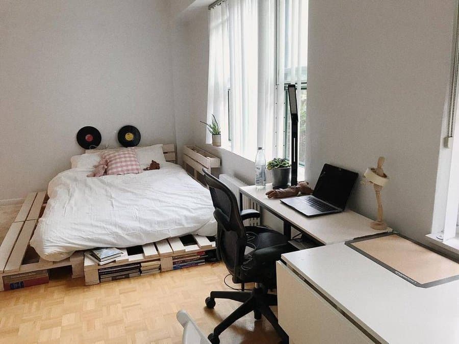 Modern minimalist bedroom with a pallet bed frame used for extra book storage, cozy white bedding, vinyl wall decor, and a workspace