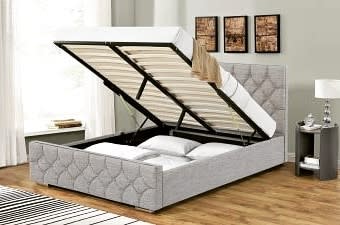 A grey upholstered storage bed with an open lift-up mattress revealing storage space underneath, in a bedroom with wooden floors