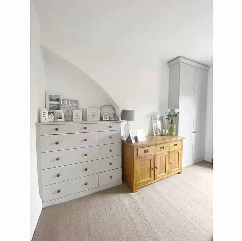 A minimalist room with a white dresser, a wooden sideboard, decor items, a lamp, a plant, and a tall cupboard against a white wall