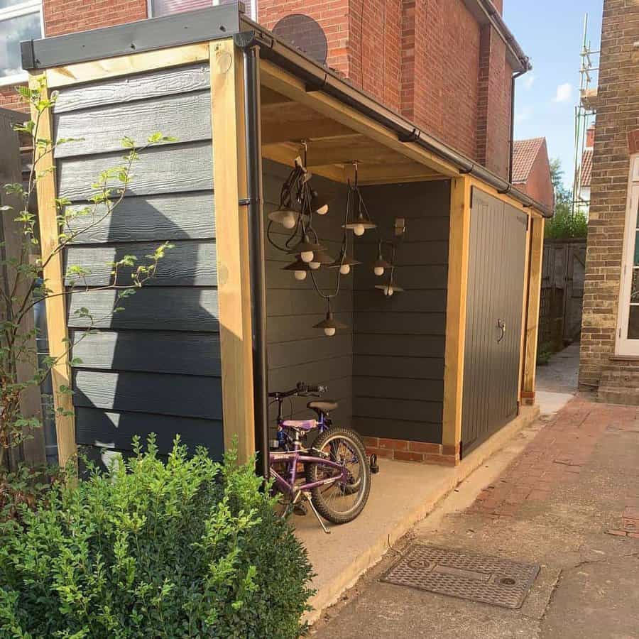 Wooden shed with dark siding, a bicycle, and hanging lights next to a brick house with a small bush in the foreground