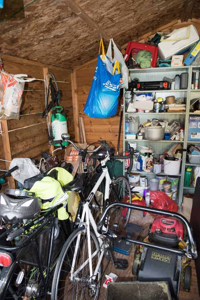 Cluttered shed with bicycles, a lawnmower, various tools, and paint cans; shelves hold assorted items and a blue bag hangs from the ceiling