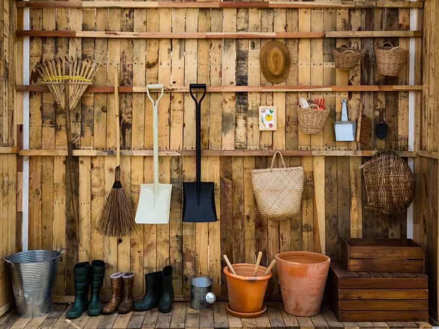 Wooden shed interior with hanging tools: rakes, shovels, baskets; buckets and boots on the floor; terracotta pots and wooden crates