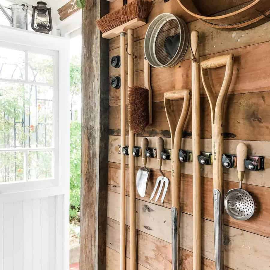 Garden tools and utensils hang neatly on a wooden wall inside a shed, next to an open white door leading outside