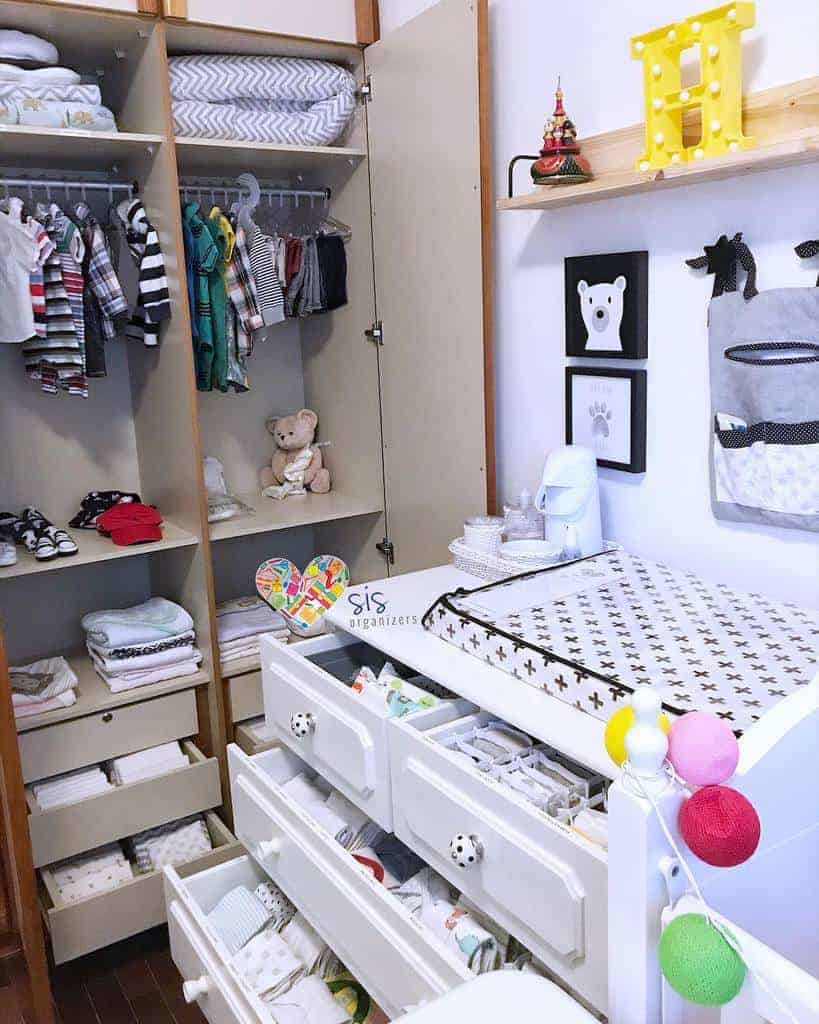 Organized nursery with a crib, changing table, open closet with baby clothes, and drawers filled with folded cloths; teddy bear on shelf