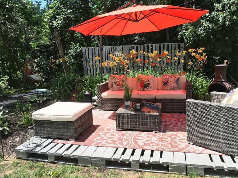 Stylish outdoor lounge with a pallet deck, rattan furniture, tropical cushions, a red umbrella, and lush greenery for a cozy backyard retreat