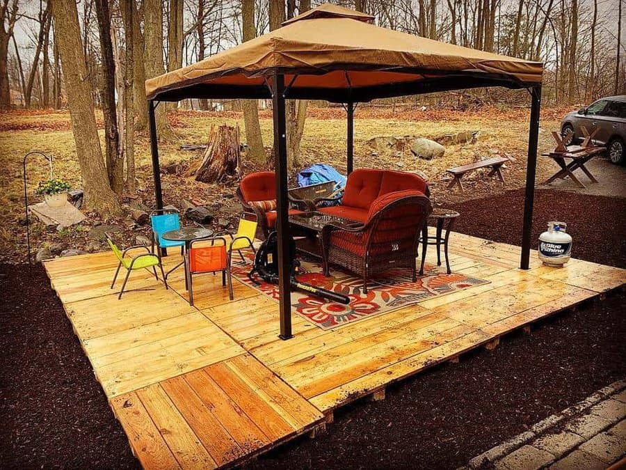 Rustic outdoor pallet deck with a gazebo, wicker seating, colorful kids' table, and a cozy rug, set in a woodland backyard setting