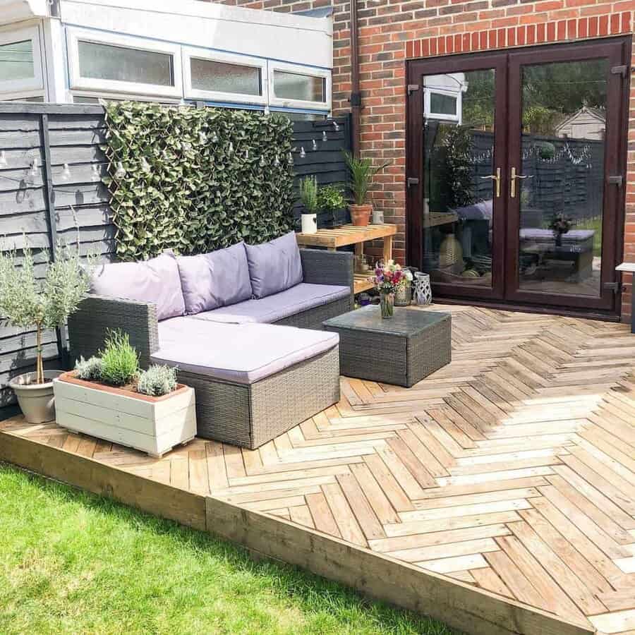 Stylish backyard pallet deck with a herringbone wood pattern, rattan seating, lavender cushions, potted plants, and a greenery wall accent