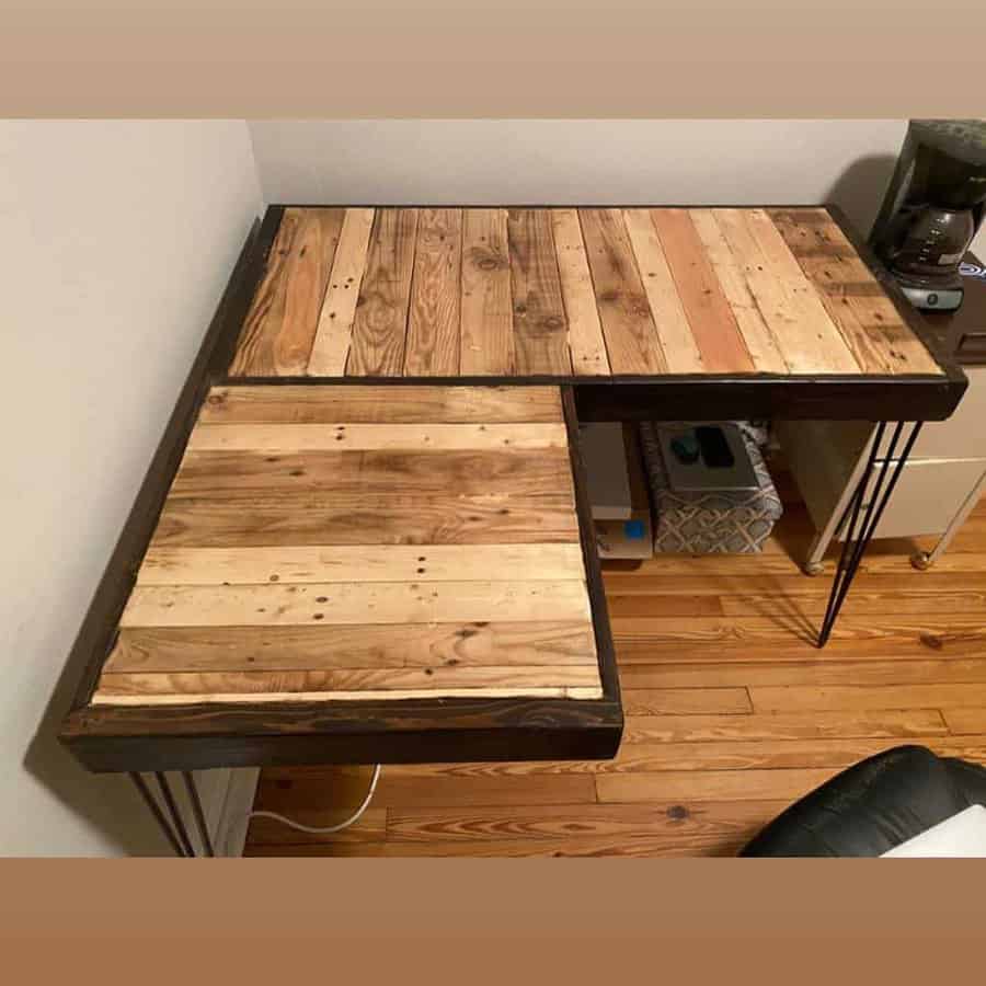 Handmade L-shaped pallet desk with a rustic wood finish, black frame, and hairpin legs, set in a cozy home office corner