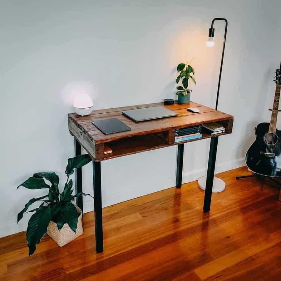 Minimalist pallet desk with built-in storage, black metal legs, warm lighting, and a sleek setup featuring a laptop, books, and plants