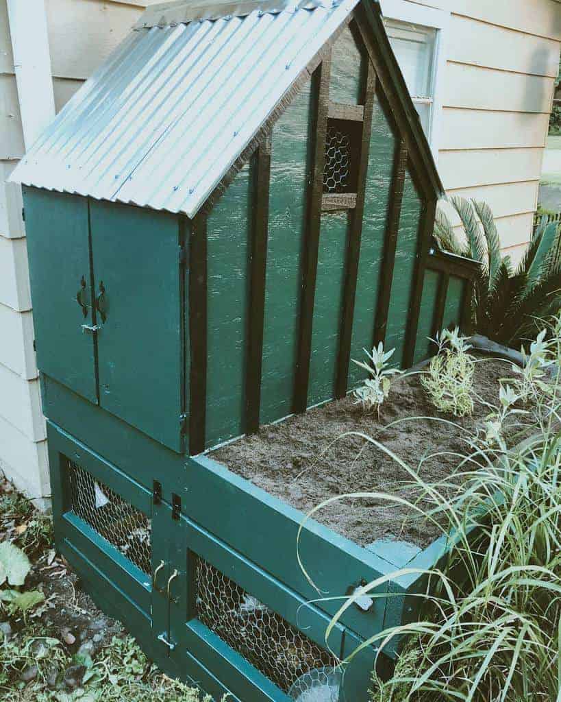 A small, green wooden chicken coop with a corrugated metal roof next to a building, featuring a planter with soil and plants on top