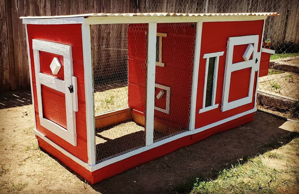 Red and white chicken coop with mesh wire, two doors, and geometric window designs, set on dirt near a wooden fence