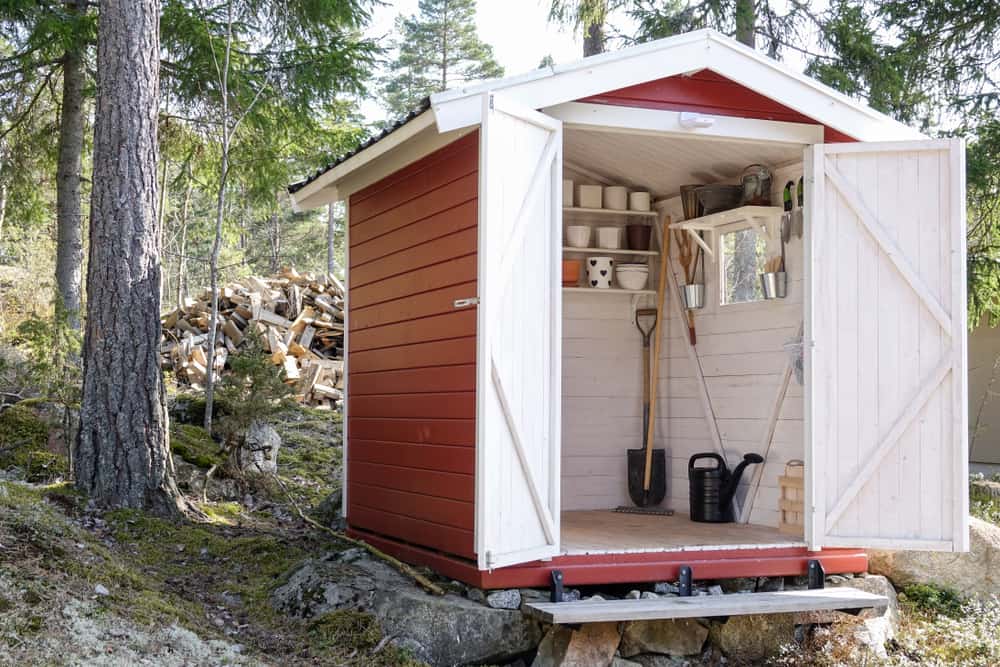 A small red and white shed in a forest, with open doors revealing gardening tools inside and a woodpile in the background