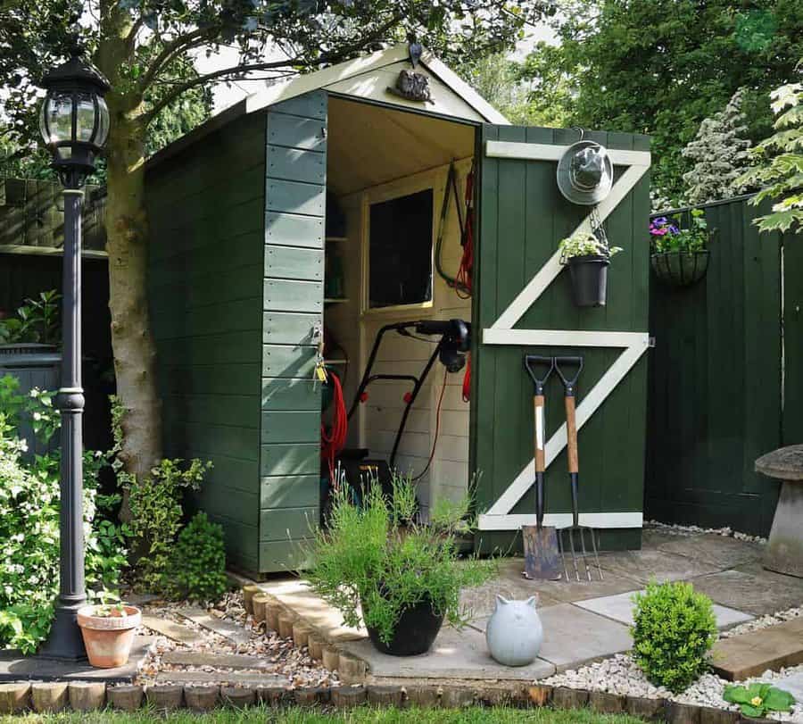 Green garden shed with white trim, surrounded by plants and tools, lantern on the left, shovel and rake propped against the wall