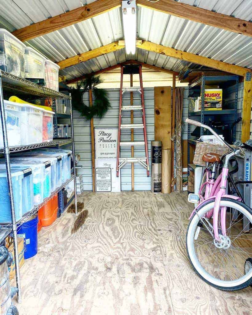 Neatly organized shed with shelves of storage boxes, a pink bicycle, ladder, and yard tools against the back wall