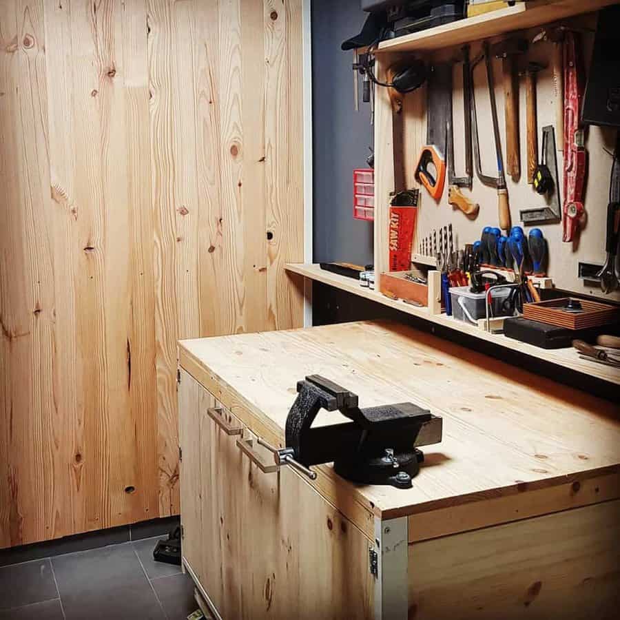 Wooden workshop with a workbench, vise, and tools like saws, hammers, and pliers on the wall; shelves hold various more items