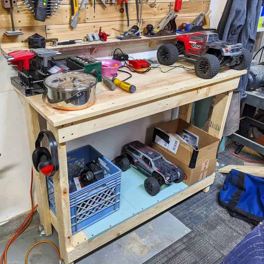 Wooden workbench with RC cars, tools, a crate, and boxes atop and below; headphones hang on the side, tool wall in the background