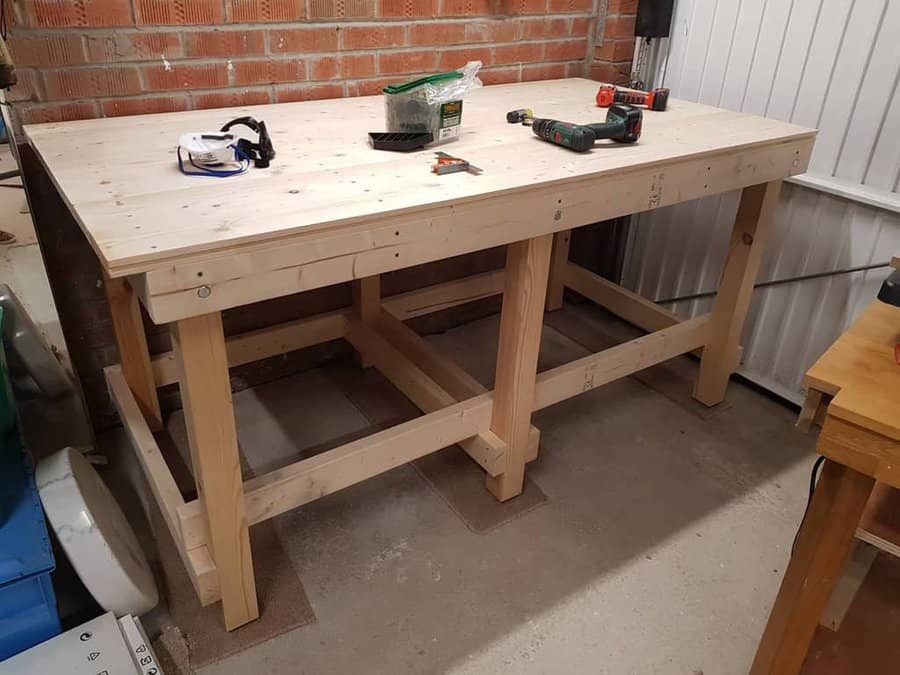 Wooden workbench in a garage with tools including a drill, mask, paintbrush, and screws on top, brick wall and white door in the background