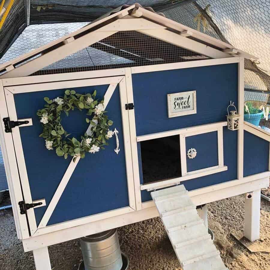 Blue and white chicken coop with a wreath and "Farm Sweet Farm" sign, featuring a small ramp and decorative lantern