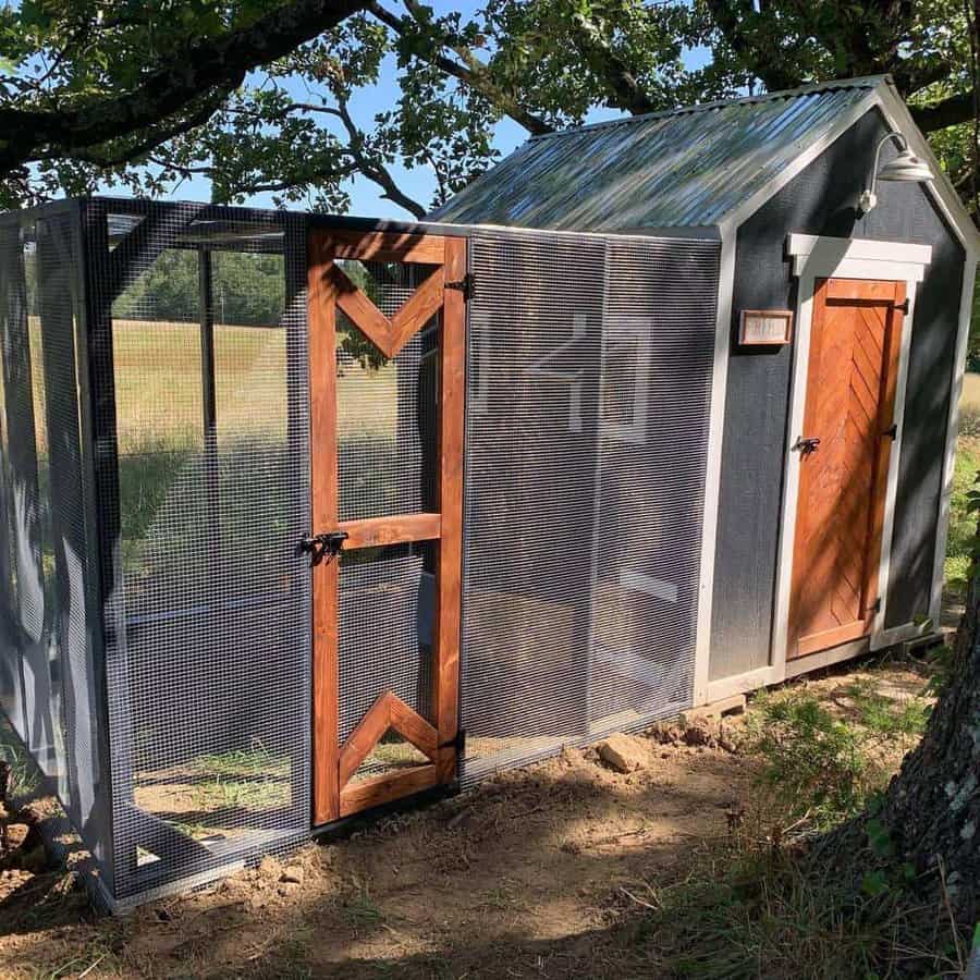 A small, gray chicken coop with a wooden door and mesh enclosure, situated under a tree in a sunny field